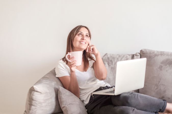 Smiling woman on sofa with a cup of coffee working on her laptop in the morning