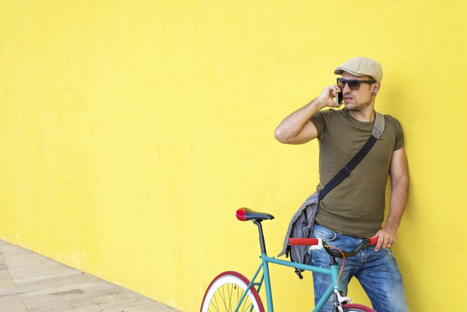 Male in hat and sunglasses standing next to yellow wall with bike and taking call on phone