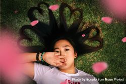 Girl with hair shaped into hearts lying on green grass with flower petal floating 5r7z24