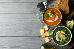 Two bowls of orange and green soup on wooden table with crackers and vegetables, copy space