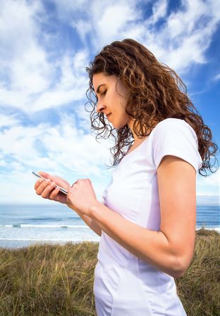 Woman in t-shirt checking phone with ocean waves in background, vertical