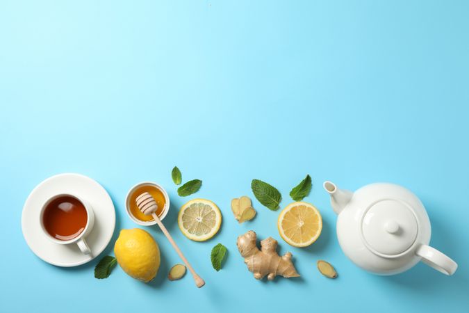 Flat lay. Cup of tea, teapot, lemon, ginger, mint, honey and dipper on blue background, copy space