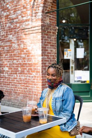 Woman sitting in front of coffee shop wearing mask smiling and looking away
