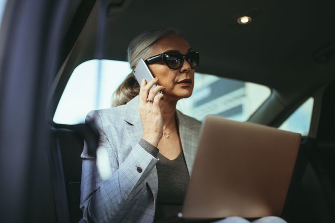 Businesswoman with laptop receiving a phone call on the backseat of a car
