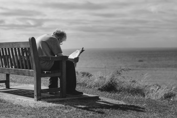 Back view of older man reading a newspaper sitting on bench at the beach in grayscale