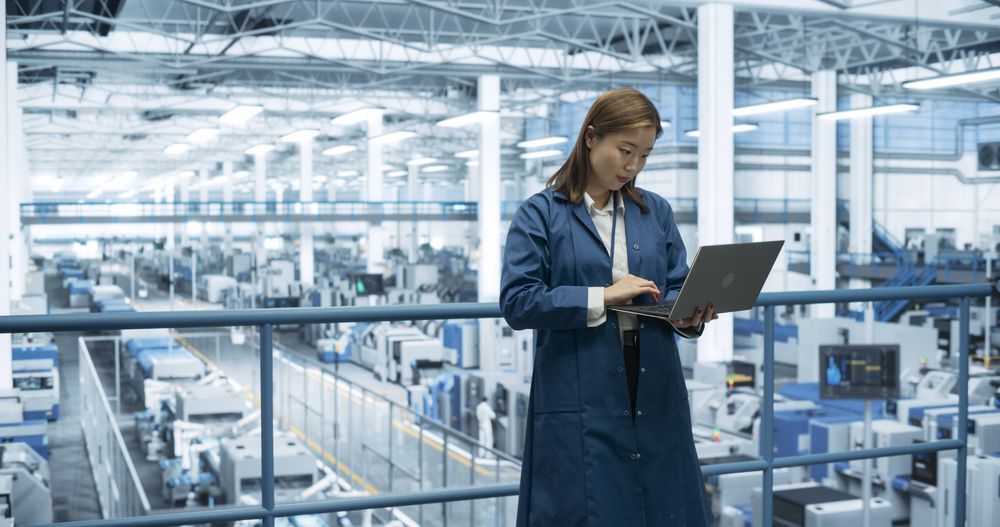 Electronics Manufacturing Technician Working on a Laptop Computer at a Factory Space. Asian Female Specialist Developing, Maintaining, Fine-Tuning Programs for Machine Production and Assembly