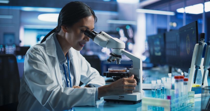 Medical Research And Development Center: Female Asian Scientist Using Microscope To Analyze Petri Dish Sample.