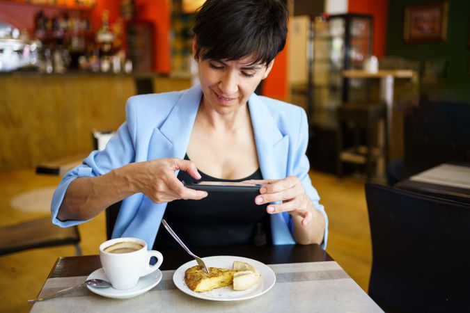 Female in trendy blue jacket sitting at table taking picture of slice of cake in cafe