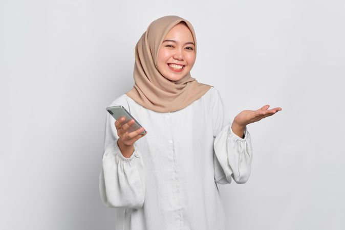 Asian female in headscarf smiling with one hand up and holding smart phone
