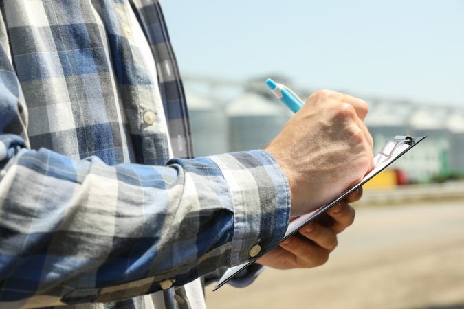 Young man with clipboard and pen against grain silos. Agriculture business