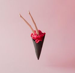 Doll legs emerging from strawberry ice cream in dark waffle cone on pink pastel background