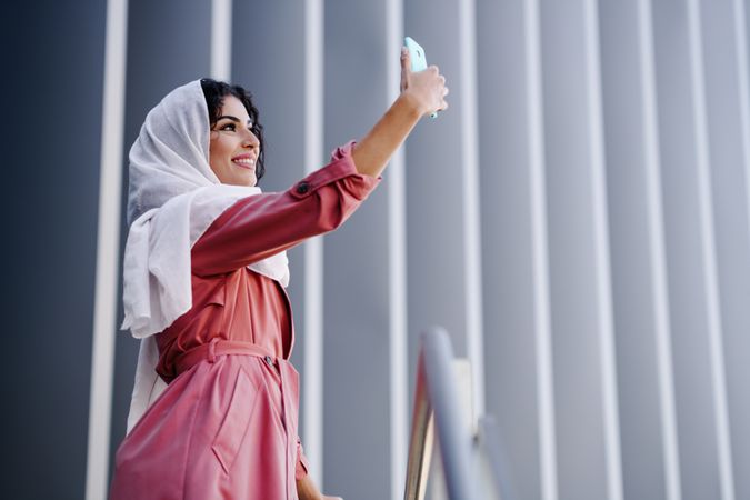 Female in headscarf and pink trench coat taking photo with her phone out