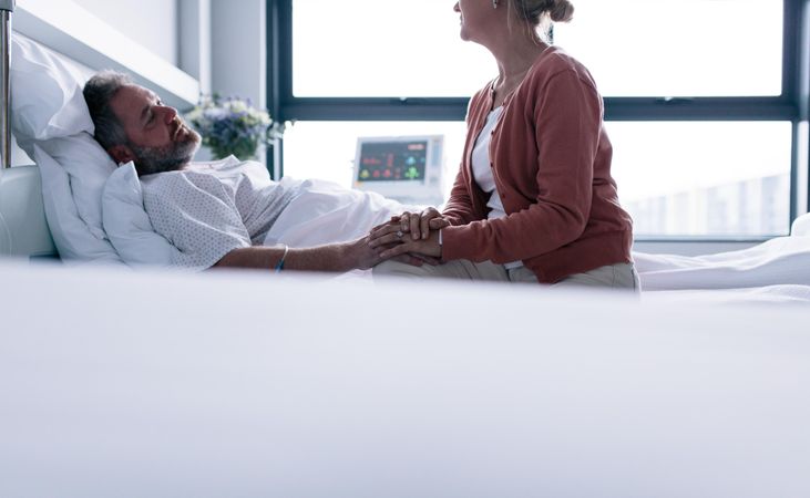 Man lying in hospital bed with woman sitting by holding hand