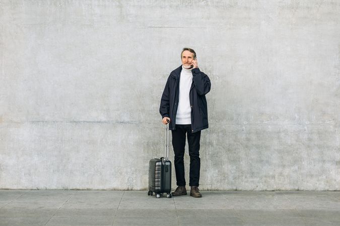 Man with suitcase outside against plain concrete wall speaking on the phone