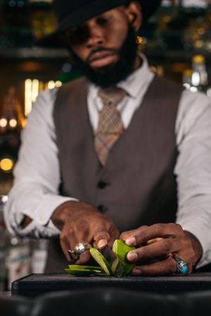 Close-up of a stylish Black bartender in a hat cutting limes to prepare cocktails