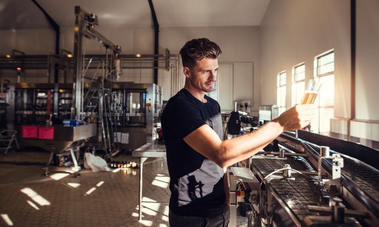 Young businessman holding sample beer glass up to examine in factory