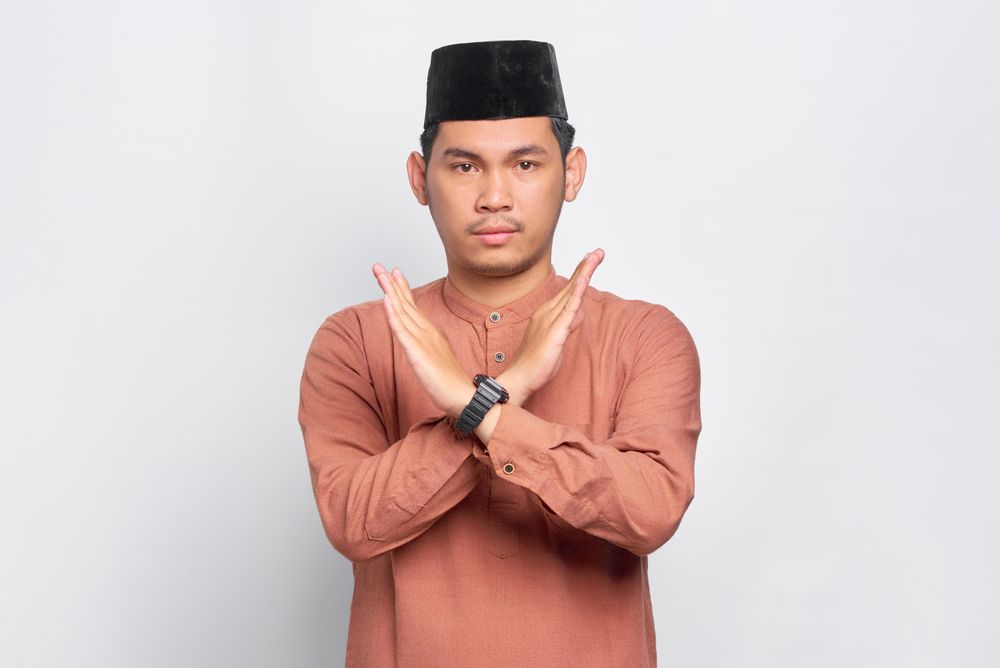 Muslim man in kufi cap make “X” sign across chest with arms