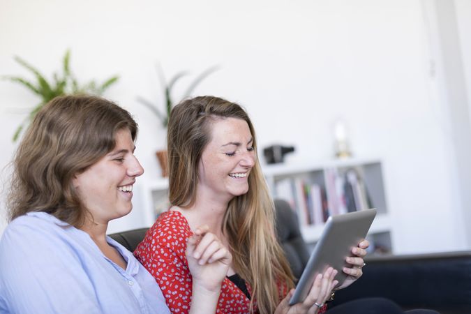 Two laughing women at home sitting on sofa using digital tablet