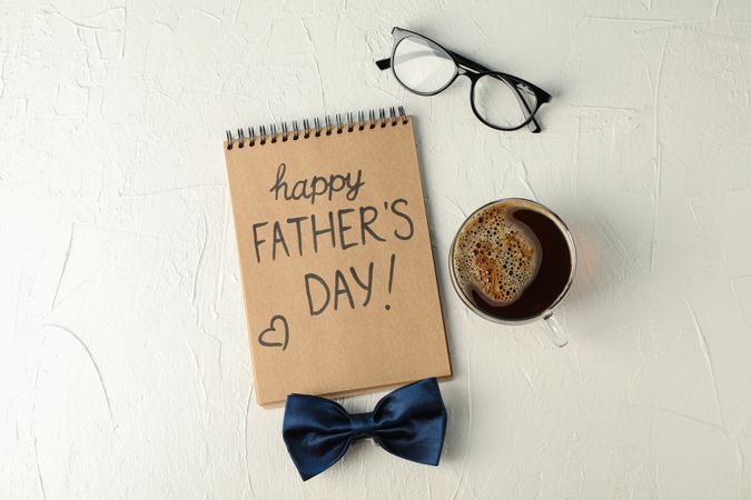 Notebook with inscription happy fathers day, blue bow tie, cup of coffee and glasses on plain background, space for text and top view