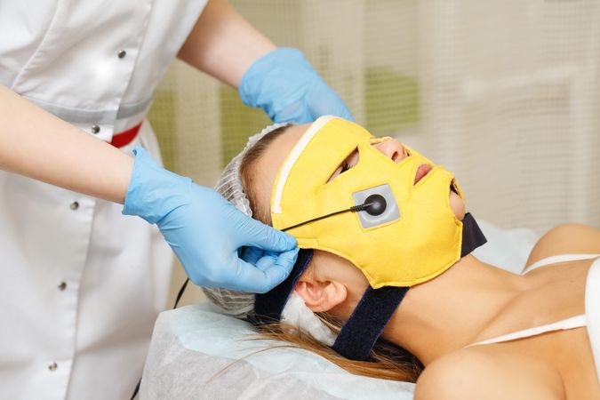 Female beautician placing yellow microcurrent mask on woman’s face during facial