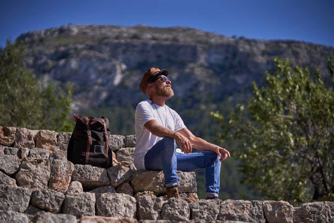Male traveler resting on stony stairs near backpack in mountains