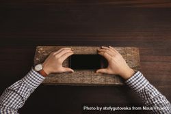 Man wearing plaid shirt and leather watch holding cell phone on slab of wood on table 49Wzm0