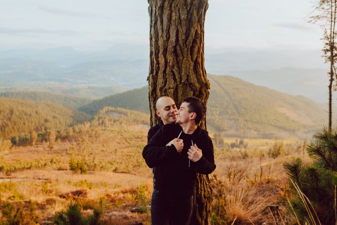 Smiling men hugging near tree in forest on hill