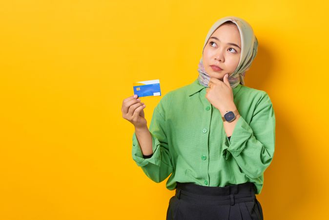 Muslim woman in headscarf and green blouse holding credit card and contemplating something