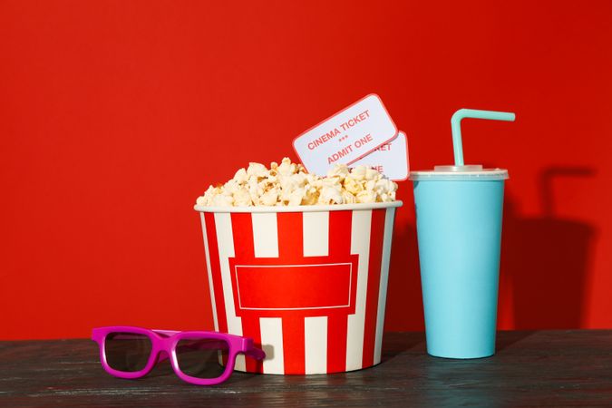 3d glasses, striped bucket with popcorn and tickets, paper cup against red background