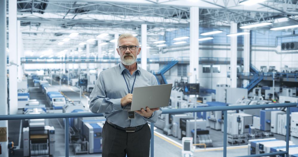 Portrait of a Handsome Mature Engineer with a Beard and Glasses Using Laptop Computer and Looking at Camera. Male Specialist Working in a Modern Electronics Factory