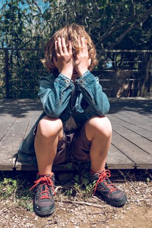 Upset boy sitting on wooden boardwalk and covering face