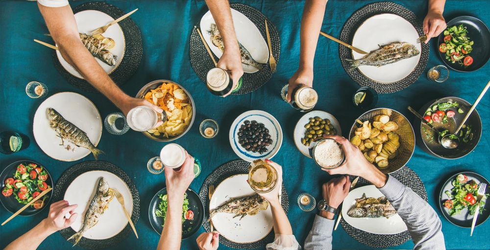 Flat-lay of human hands toasting with beer glasses over grilled sea bass, wide composition