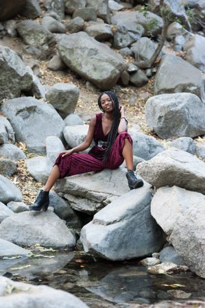 Woman in velvet jumpsuit and black boots sitting on boulders with hand by her neck