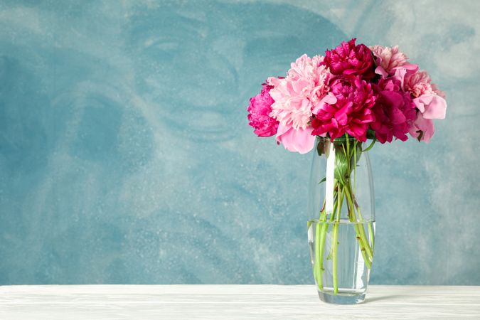 Glass vase with bouquet of beautiful peonies on table against blue background, space for text
