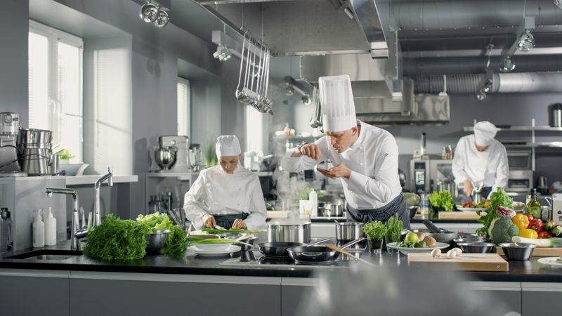 Famous Chef Works in a Big Restaurant Kitchen with His Help. Kitchen is Full of Food, Vegetables and Boiling Dishes. He is trying taste.