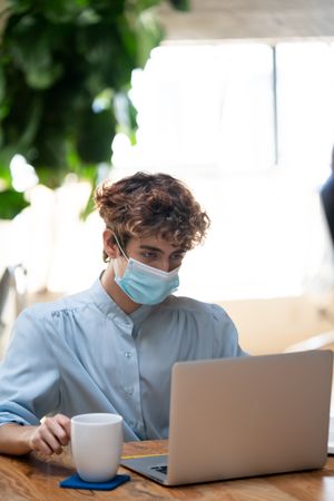 Nonbinary person in medical face mask working on a laptop with a cup of coffee