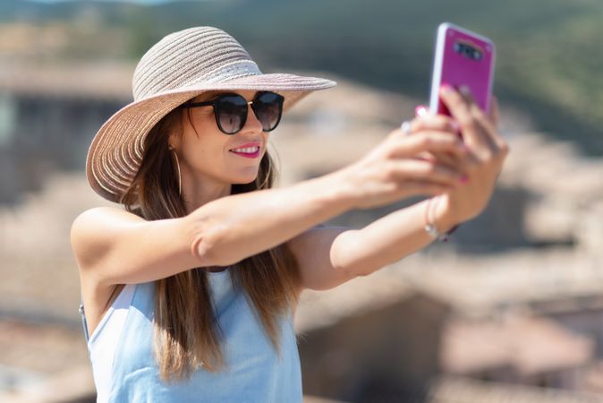 Positive woman in hat taking selfie on smartphone while standing on street