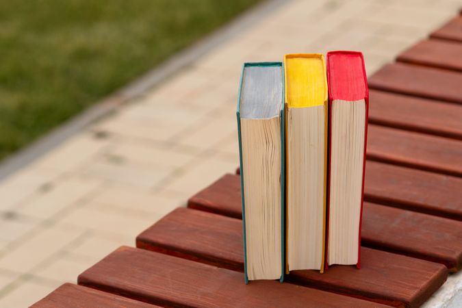 Three hardcover books vertically placed on wooden park bench.