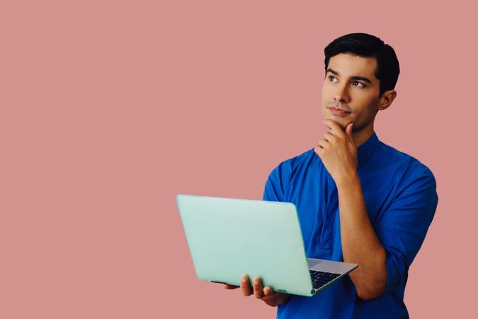 Contemplative Latino man looking up while holding laptop in pink room, copy space
