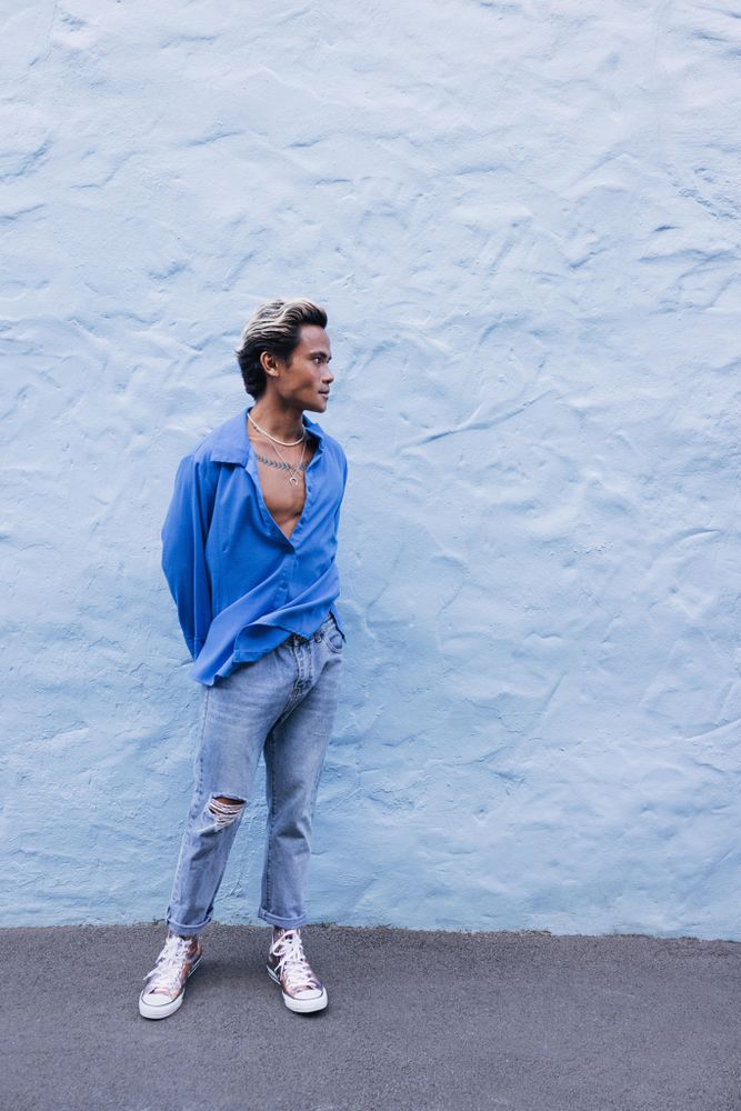 Asian male standing in front of wall in blue shirt and jeans, vertical