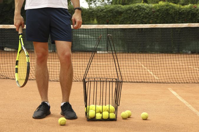 Man hold racket on clay court with basket of tennis balls