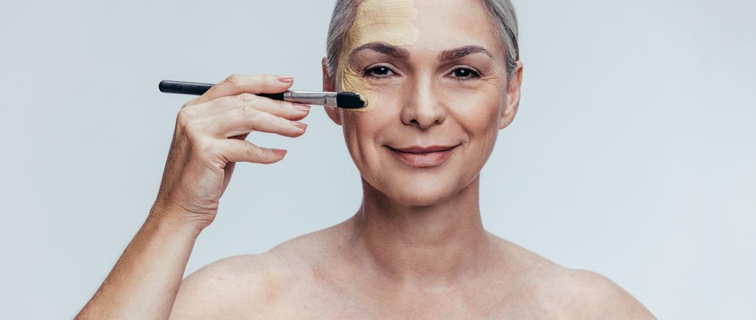 Beautiful woman with gray hair applying foundation on face with makeup brush
