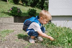 Adorable child sitting with backpack on ground and playing with plants