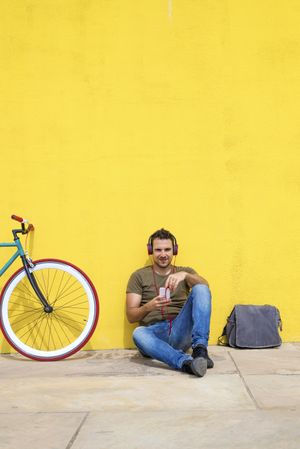 Male sitting in front of yellow wall next to bike and listening to music on smartphone, vertical