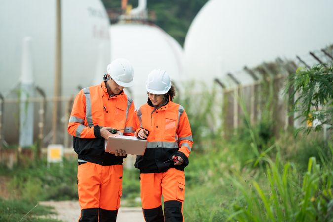 Two engineers in safety gear talking work near spherical gas storage tanks. woman is pointing forward while the man checks a laptop, indicating active discussion and site coordination in an industrial