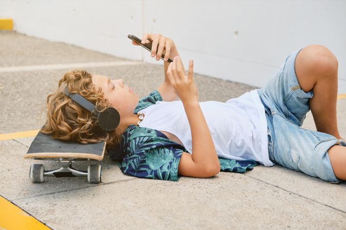 Boy lying on skateboard on street and using smartphone