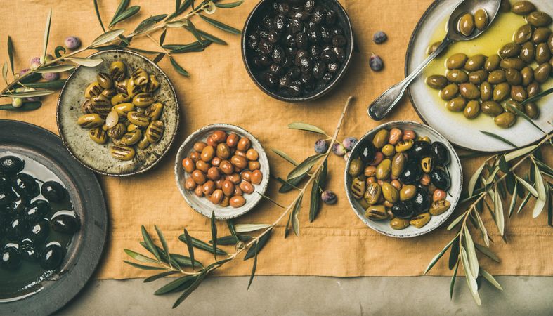 Olives in bowls with branches, on beige table linen, with serving spoon, wide composition