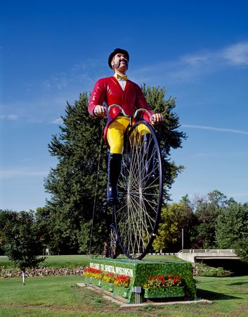 Statue of man in red coat, yellow pants, a curled mustache on 30’ bicycle, Sparta, Wisconsin