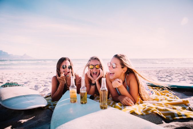 Group of young women sitting in the sand with a surfboard and beer