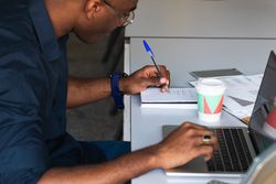 Black male writing in notepad near laptop in office